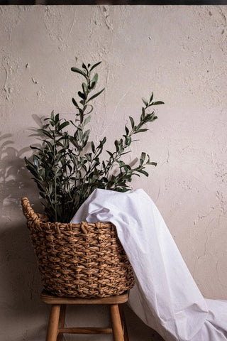 White stonewashed cotton curtain fabric folded in a basket with olive branches. Handwoven in Denizli, OEKO-TEX. Vivamaison.
