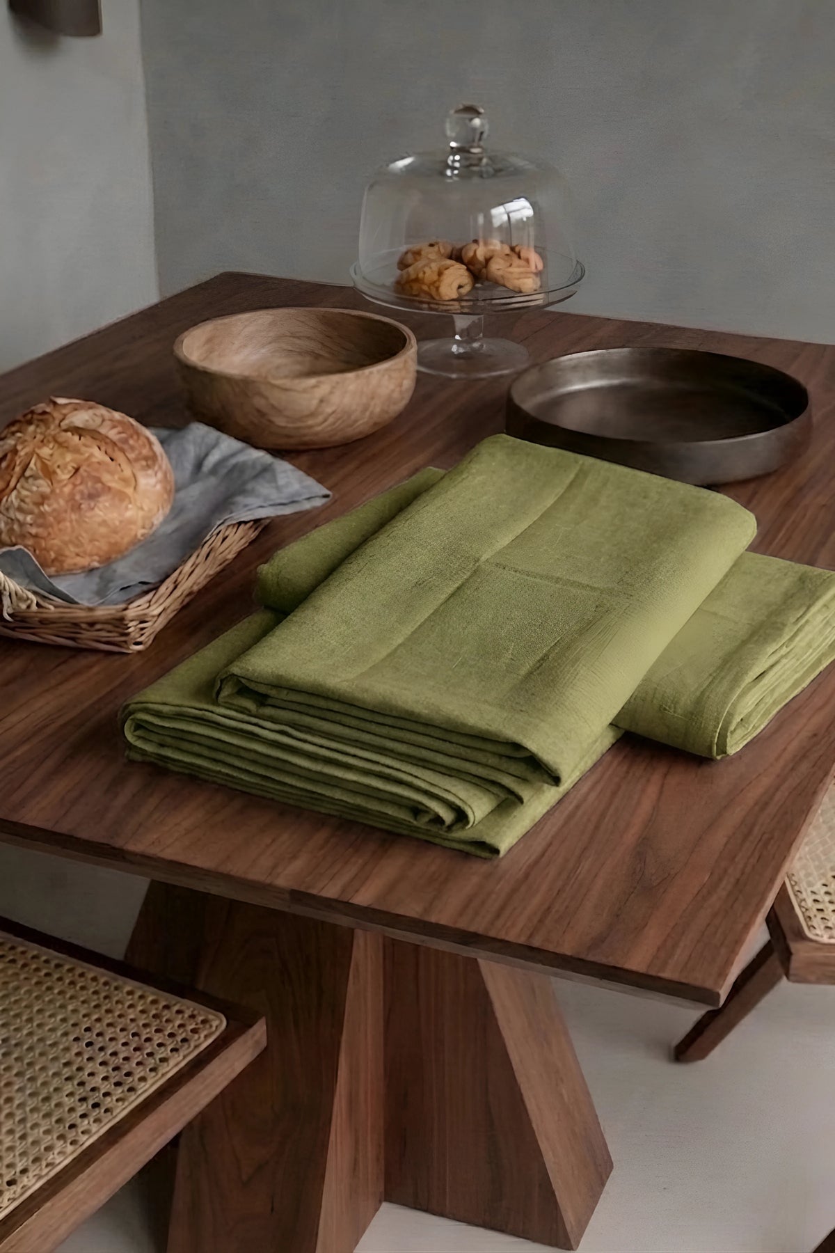 Olive Green plain linen cotton blend tablecloth folded on a walnut dining table beside a bread basket. 9 sizes, handmade in Denizli, OEKO-TEX. VivaMaison.
